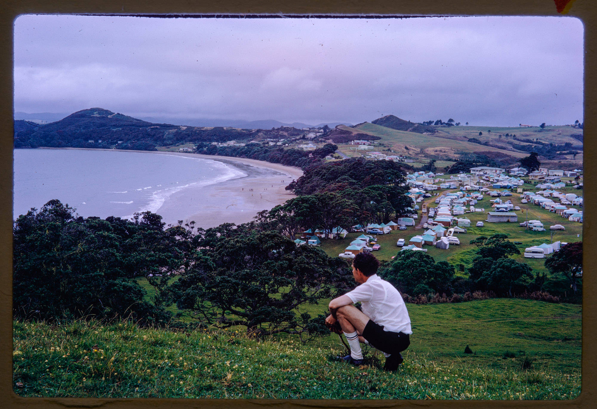 Keith Thomson kneeling on a hill above Coopers Beach, with Coopers Beach Motor Camp in the valley below.